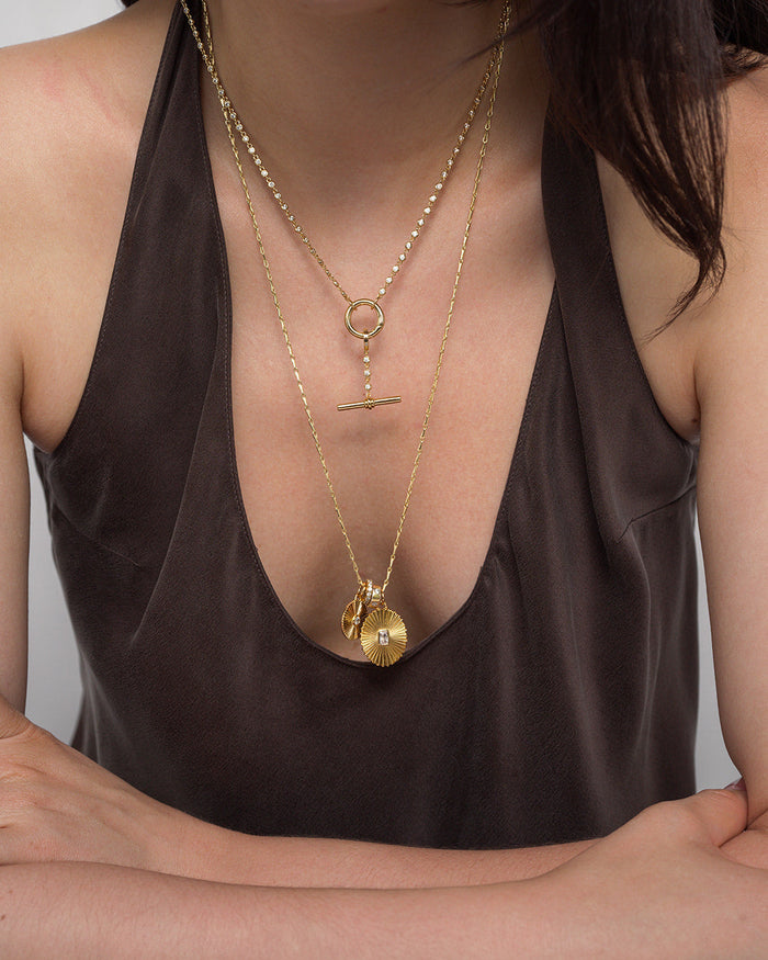A woman in a dark, sleeveless top rests her arms on a surface, showcasing layered Mod + Jo 14k gold necklaces featuring an emerald-cut crystal and the Cora Fluted Charm. The cropped image highlights her upper torso and jewelry.
