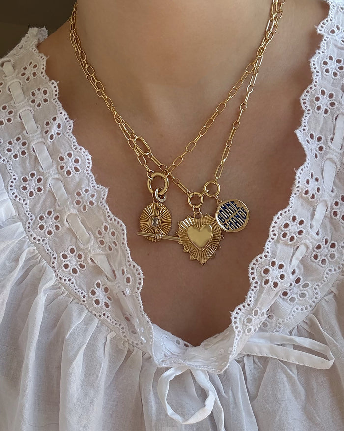 A person wearing a white eyelet lace blouse and two 14k gold chain necklaces with three gold charms: a fan, a heart, and a round charm inscribed with "MAMA CERAMIC." One necklace showcases the elegant Mod + Jo Cora Fluted Charm, enhancing the timeless allure of their jewelry ensemble.
