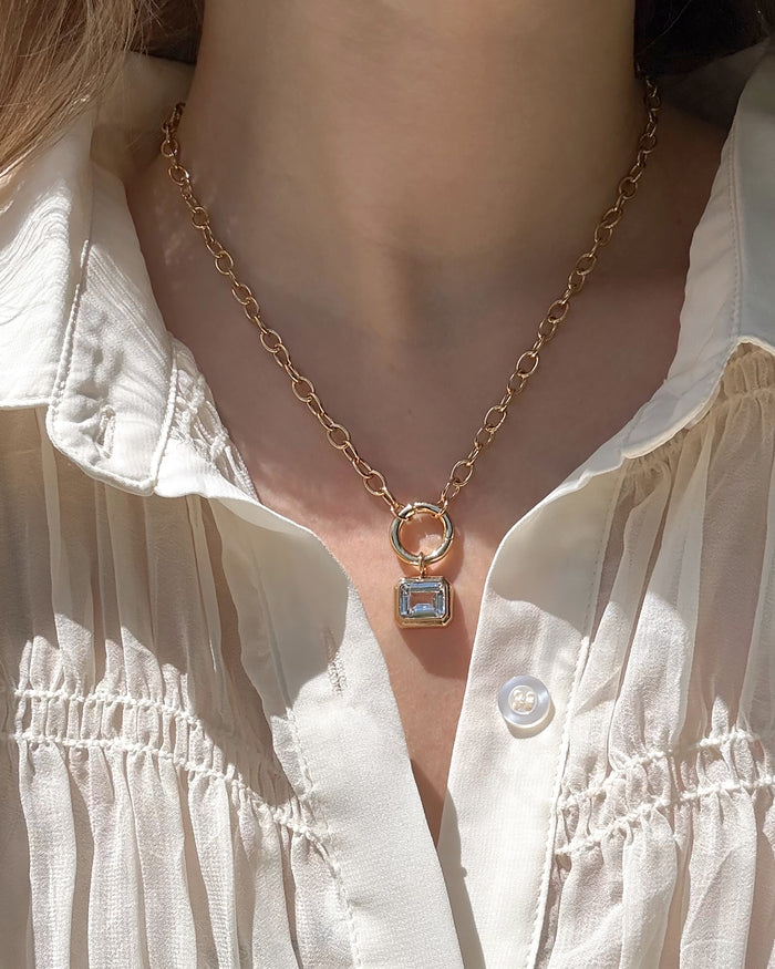 A close-up of a person wearing a cream pleated blouse and the Mod + Jo Darcy Paperclip Enhancer Chain, a gold 14k plated necklace with a rectangular pendant featuring a clear gemstone.