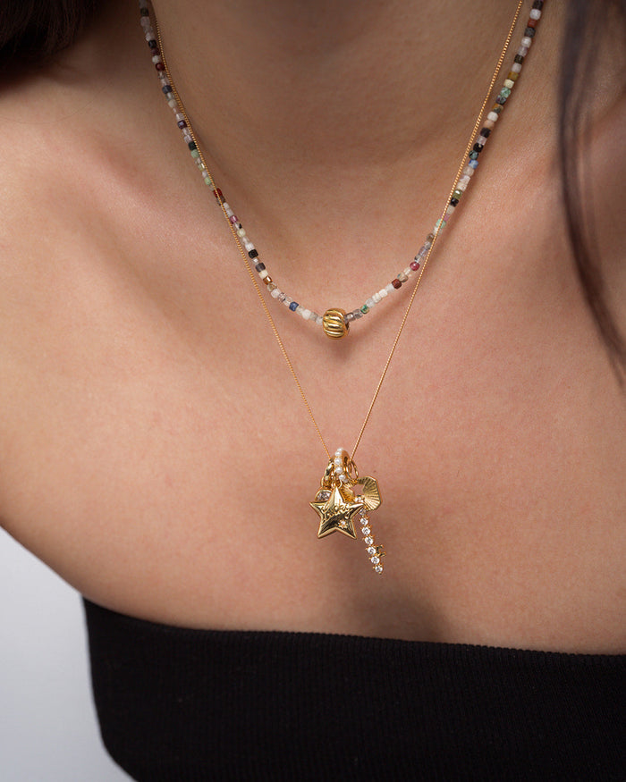 A woman wearing two layered necklaces from Mod + Jo: one adorned with colorful beads and a gold bead, and the other featuring the Elara Lucky Star Charm in 14k gold, along with heart and crystal pendants—styled over a black strapless top.
