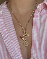 A person wearing a pink and white striped shirt and a gold necklace adorned with charms, including a key, padlock, the Mod + Jo Elara Lucky Star Charm, and a round Louis Vuitton pendant. The necklace showcases elegant 14k gold jewelry details for a chic finishing touch.