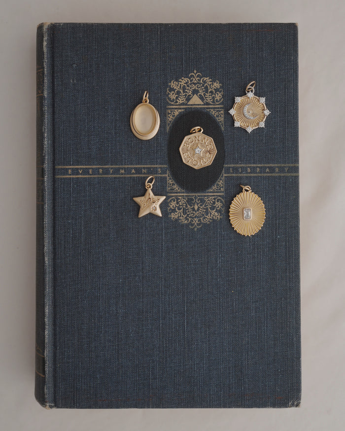 A dark blue vintage book with gold detailing lies on a white surface. Five Rory Gemstone Charms by Mod + Jo, each featuring unique shapes, intricate designs, and shimmering Moonstone accents in gold and silver tones, are arranged on the book’s cover.
