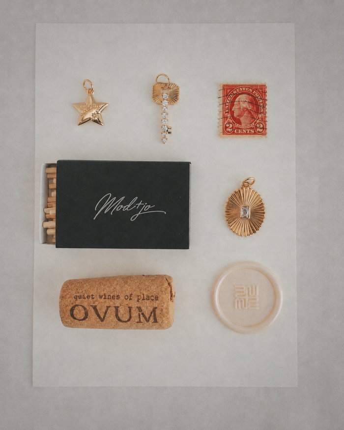 A flat lay showcases 14k gold jewelry by Mod + Jo, including a gold star necklace with the Elara Lucky Star Charm, displayed alongside a red stamp, a black matchbox, a cork with "OVUM," and a round translucent object, all neatly arranged on white paper.
