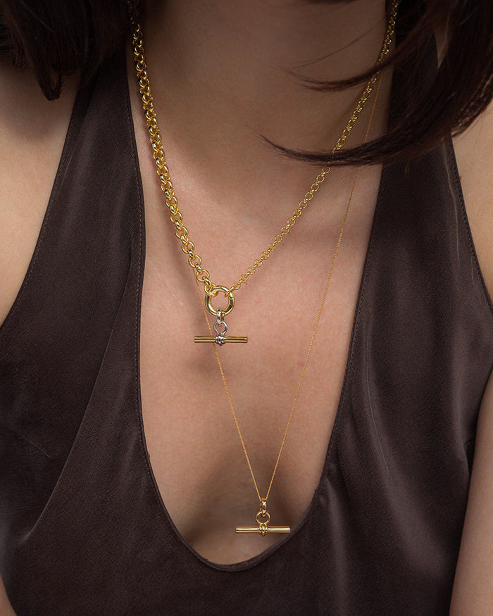 A person wearing a dark, deep V-neck top showcases two layered Mod + Jo Elliot Toggle Charm necklaces in 14k gold, each featuring a horizontal bar pendant and distinct chain styles, set against their bare upper chest.