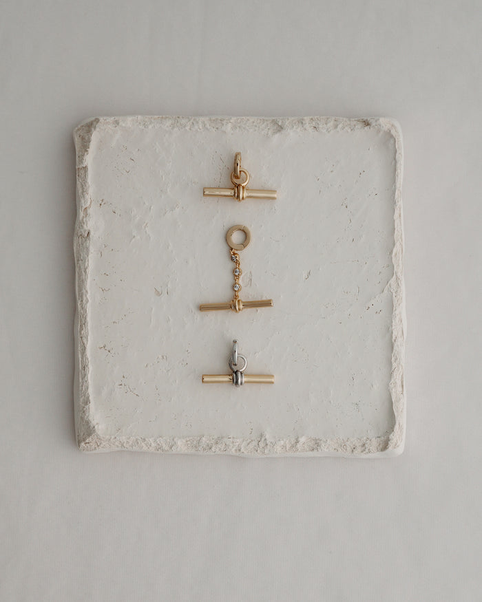 Three metal toggle clasps—two in gold and one in silver—are arranged vertically on a square, textured white stone tile set against a light background, echoing the elegant detail of Mod + Jo's Elliot Toggle Charm and the refined style characteristic of their 14k gold-inspired jewelry.