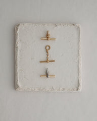 Three metal toggle clasps—two in gold and one in silver—are arranged vertically on a square, textured white stone tile set against a light background, echoing the elegant detail of Mod + Jo's Elliot Toggle Charm and the refined style characteristic of their 14k gold-inspired jewelry.