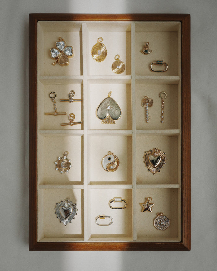 Showcased in natural light, a wooden jewelry box with 15 beige sections displays an array of gold and silver charms and earrings—including the Mod + Jo Elliot Toggle Charm, 14k gold jewelry, hearts, stars, eyes, and other unique architectural designs.