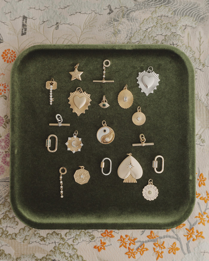 Nora Lock Carabiner charms by Mod + Jo—crafted in gold and silver finishes, featuring a range of shapes like hearts, stars, a yin-yang motif, and geometric forms (some embellished with natural semi-precious stones such as blue lace agate and highlighted with elegant 14k plating)—are artfully arranged on a dark green velvet tray. The backdrop is a richly patterned floral fabric.