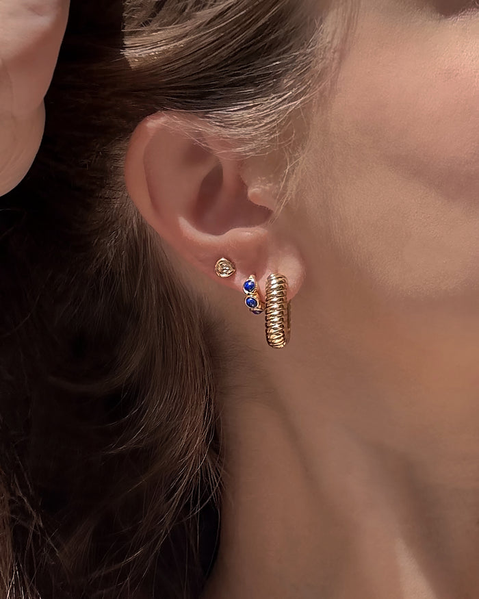 A close-up of a woman’s ear showcases three gold earrings: a simple stud, a stud featuring two blue stones, and the chunky textured Elly French Rope Hoop by Mod + Jo. Her brown hair is tucked behind her ear, drawing attention to the elegant details of her Mod + Jo gold hoop earring.
