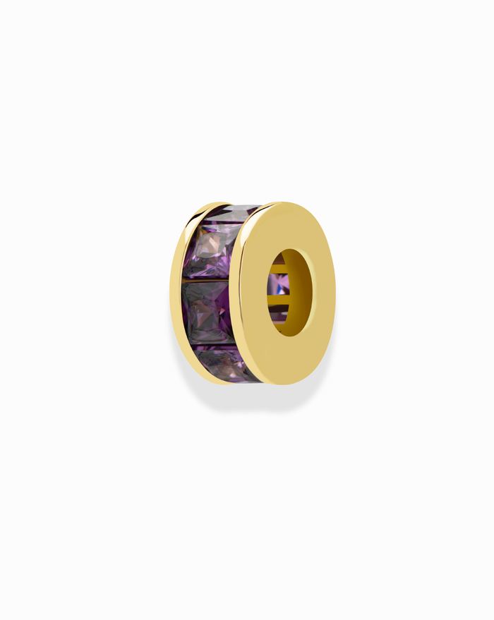 The Mod + Jo Este Birthstone Charm Spacer is a gold bead with a hollow center, featuring a row of rectangular purple gemstones around the edge—ideal for personalizing necklaces. Shown on a plain white background.