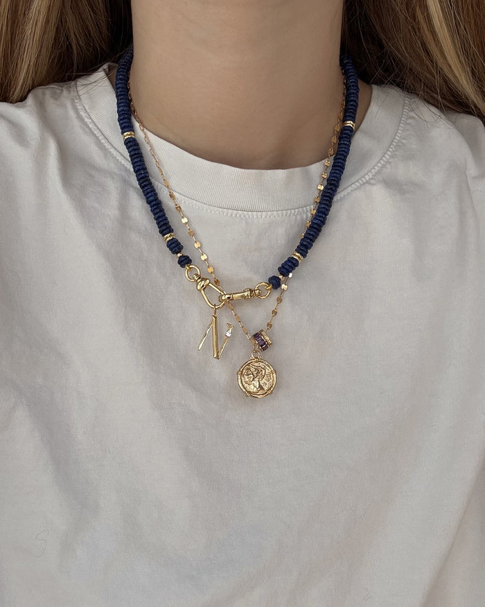 A person wearing a white shirt with three layered necklaces: one gold plated letter charm necklace, one with dark blue beads, and the Mod + Jo Este Birthstone Charm Spacer—a chic blend of personalized accessories.