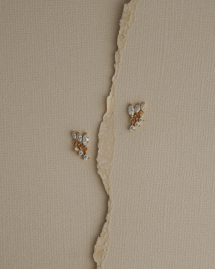 Two Gabby Marquise Dangle Studs by Mod + Jo, gold plated and adorned with sparkling cubic zirconia, rest on a textured beige surface, separated by a torn vertical strip of paper.