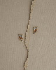 Two Gabby Marquise Dangle Studs by Mod + Jo, gold plated and adorned with sparkling cubic zirconia, rest on a textured beige surface, separated by a torn vertical strip of paper.