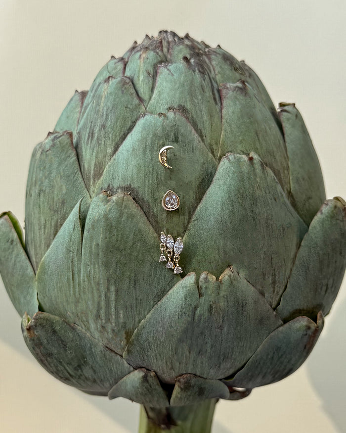 A green artichoke displays three Mod + Jo Gabby Marquise Dangle Studs—each featuring a gold plated crescent moon, a round cubic zirconia gem, and a butterfly—arranged vertically on its front against a plain background.