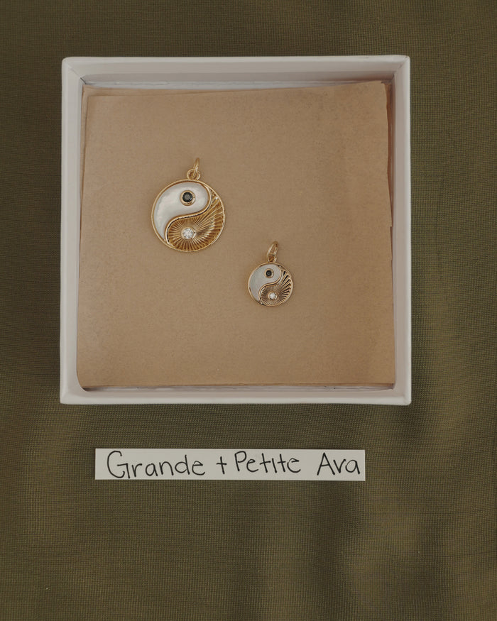 Two Mod + Jo Grande Ava Yin Yang charms, one large and one small 14k gold pendant, are displayed in a box on brown paper. Below the box is a label that reads "Grande + Petite Ava" on a green fabric background.