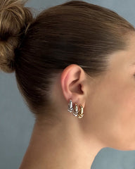 A woman with brown hair in a bun wears three small hoop earrings—including Mod + Jo’s Grande Levi Huggies in gold—on her left ear, alongside silver and rose gold hoops. She is facing right against a plain, soft blue background.