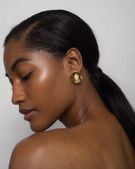 A woman with smooth, glowing skin and straight dark hair in a low ponytail is shown in profile, wearing Mod + Jo's Hunter Statement Studs—bold blue chalcedony and 14k gold earrings—against a plain light background.