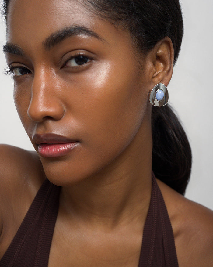 A woman with smooth, glowing skin and straight dark hair pulled back wears a sleeveless brown top and Mod + Jo's Hunter Statement Studs—large earrings with a luminous mother of pearl center—looking confidently at the camera against a plain background.