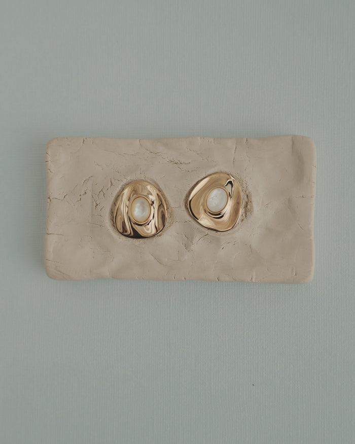The Mod + Jo Hunter Statement Studs, featuring 14k gold-toned irregular shapes with pearl centers, are shown on a textured beige clay slab, set against a light blue background.