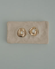 The Mod + Jo Hunter Statement Studs, featuring 14k gold-toned irregular shapes with pearl centers, are shown on a textured beige clay slab, set against a light blue background.
