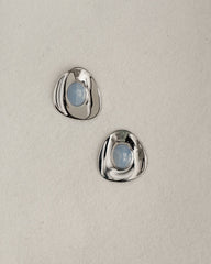 The Mod + Jo Hunter Statement Studs are two shiny, irregular silver earrings with round blue chalcedony centers, displayed on a light beige textured surface.