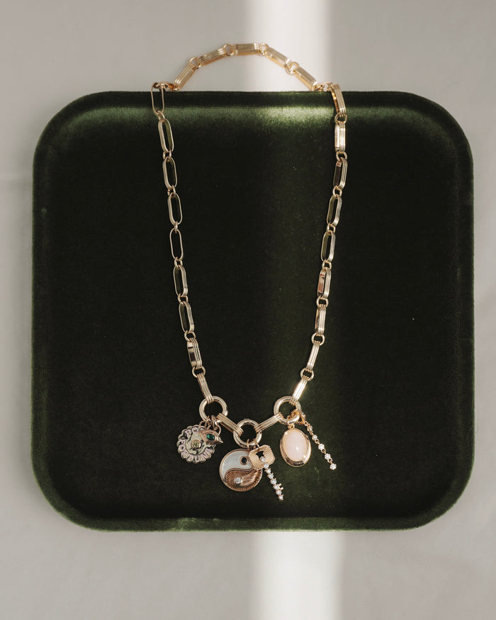 The Jagger Charm Clip Necklace by Mod + Jo, crafted in 14k gold and featuring four unique charms—a yin-yang, a flower, an oval stone, and a petite hanging pendant—is elegantly displayed on a dark green velvet tray with sunlight creating gentle shadows.