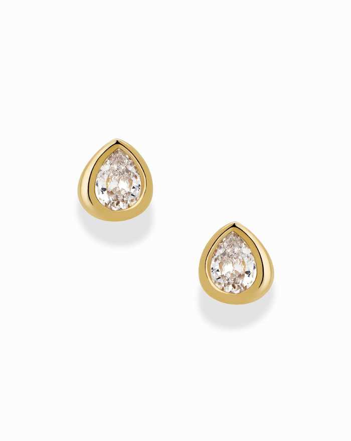 The Joni Bezel Studs by Mod + Jo are gold-plated earrings, each showcasing a clear, pear-shaped cubic zirconia gemstone in a simple bezel setting and displayed on a white background.