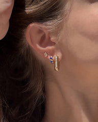 A close-up of a woman’s ear showcases three gold plated earrings from Mod + Jo: a small stud, the Joni Bezel Studs featuring double blue gems, and a textured gold hoop. Her brown hair is neatly tucked behind her ear.