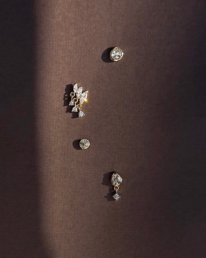 Four unique earrings from Mod + Jo, including the Joni Bezel Studs, are arranged vertically on a dark brown background. They showcase gold- and silver-toned designs in various shapes with sparkling stones like cubic zirconia, all casting soft shadows.
