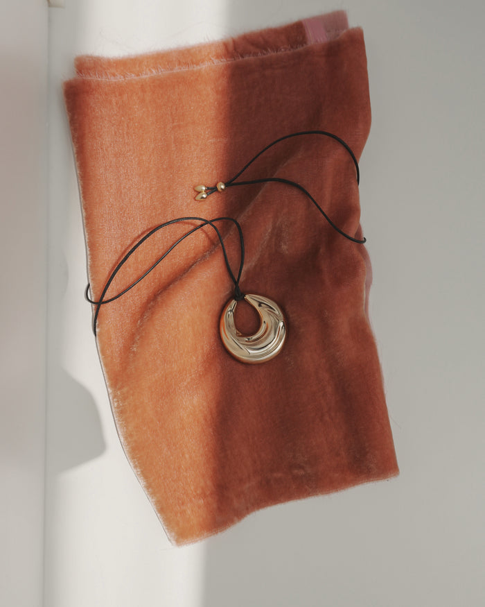 The Kira Cord Necklace by Mod + Jo features a gold pendant on a cord, displayed on soft burnt orange fabric as sunlight creates gentle shadows across the scene.