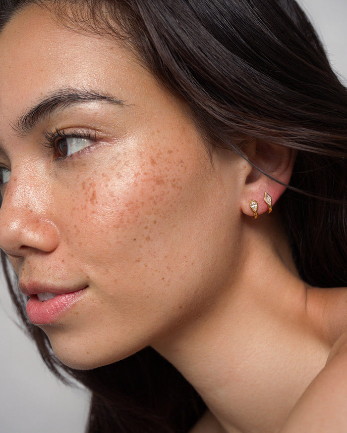 A woman with smooth, freckled skin and long dark hair wears two small gold-plated Leighton Teardrop Huggies by Mod + Jo. She gazes slightly to the left with a soft, natural expression against a plain background.