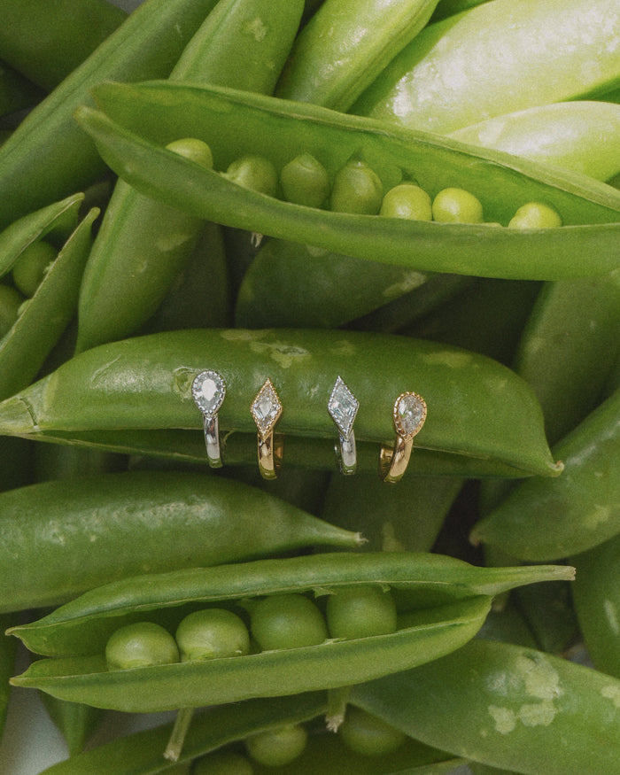 Four Leighton Teardrop Huggies by Mod + Jo are displayed on opened pea pods, surrounded by fresh green peas and pods, their brilliant stones sparkling under the light.