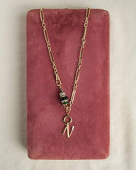 The Mod + Jo Liam Space Bar Chain Necklace features 14k gold plating, a cluster of black, clear, and green beads, and a gold letter N charm, displayed on a pink velvet rectangular box.