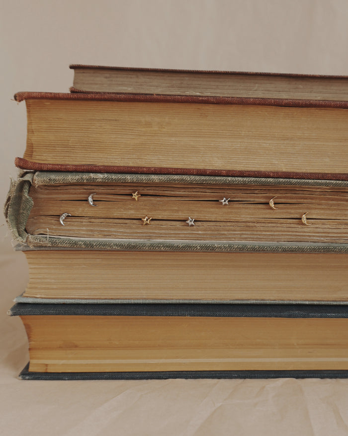 A stack of four old books lies horizontally, with Mod + Jo’s Archie Moon Studs inserted along the edges of the pages in the second book from the top. The background is neutral and plain.