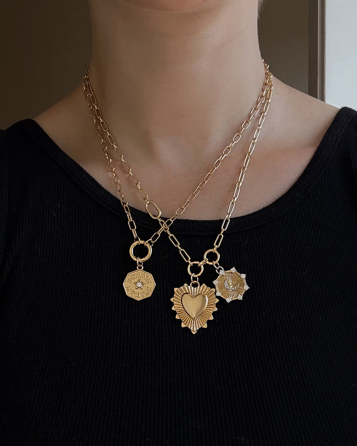 A person wearing a black ribbed top and two gold chain necklaces, each featuring ornate gold pendants in various shapes—including a heart, geometric designs, and the Lyra Moon Charm by Mod + Jo inspired by celestial jewelry.