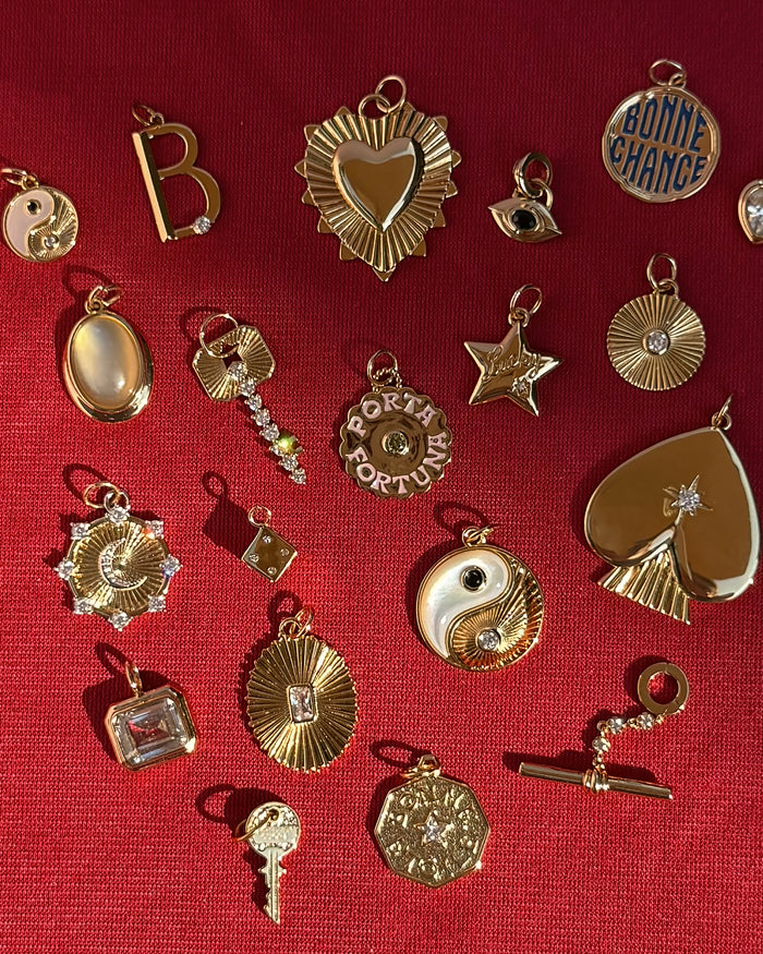 Scattered on a red textured surface are gold and jeweled charms in various shapes—hearts, stars, keys, yin-yang symbols—and the Lyra Moon Charm by Mod + Jo, whose delicate crescent evokes celestial jewelry. Some charms are engraved with phrases like “BONNE CHANCE” and “PORTA FORTUNA.”.