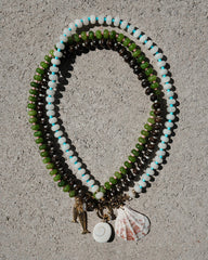 Three Mod + Jo Macey Gemstone Necklaces, arranged in ovals on a gray textured surface, feature genuine gemstones and pendants—a seahorse, spiral shell, and scallop shell. One design is accented with elegant white jade beads.