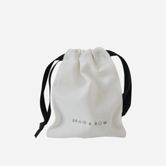 A small white drawstring bag with black ribbon handles and BRAID & BOW printed on the front, perfect for gifting the Medium Caramel Swirl Hair Claw or another secure-hold hair accessory.
