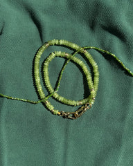 The Merit Gemstone Necklace by Mod + Jo, featuring green beads, gold accents, and a 14k gold-filled clasp, is elegantly displayed coiled on textured green fabric.