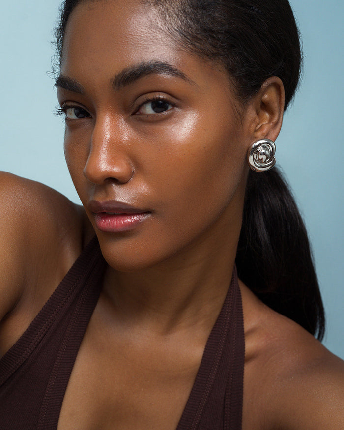 A woman with smooth dark skin and straight dark hair pulled back wears Milley Spiral Statement Studs by Mod + Jo and a brown halter top, posing confidently against a light blue background.
