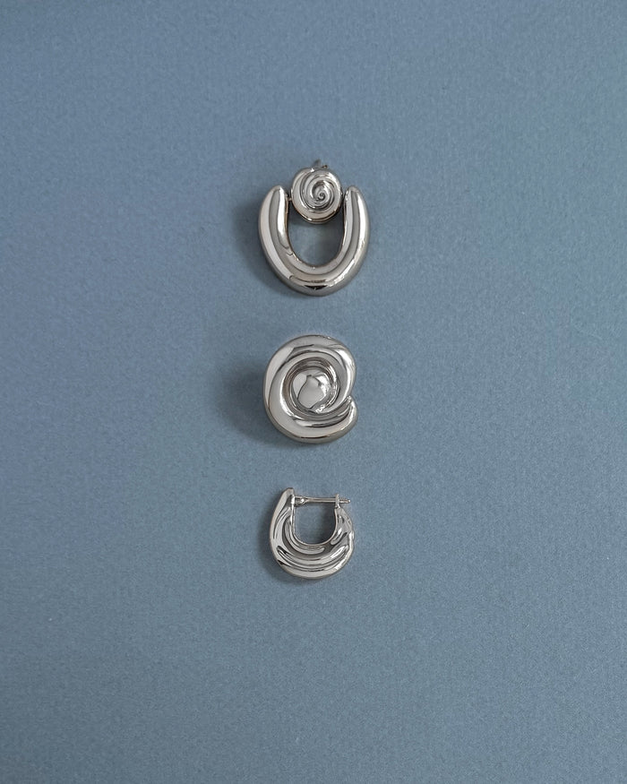 Three silver earrings from Mod + Jo are arranged vertically on a light blue textured background. The top and bottom are crescent-shaped, while the center features the Milley Spiral Statement Studs—an elegant hypoallergenic choice.