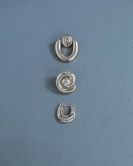 Three silver earrings from Mod + Jo are arranged vertically on a light blue textured background. The top and bottom are crescent-shaped, while the center features the Milley Spiral Statement Studs—an elegant hypoallergenic choice.