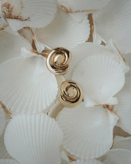 The Milley Spiral Statement Studs by Mod + Jo, crafted in shiny 14k gold, are displayed atop overlapping white scallop seashells with light brown edges.