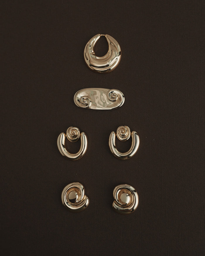 Five pairs of Mod + Jo Milley Spiral Statement Studs in 14k gold are artfully arranged on a dark brown background—ideal for daily wear or to add a unique, abstract touch with their swirling design.