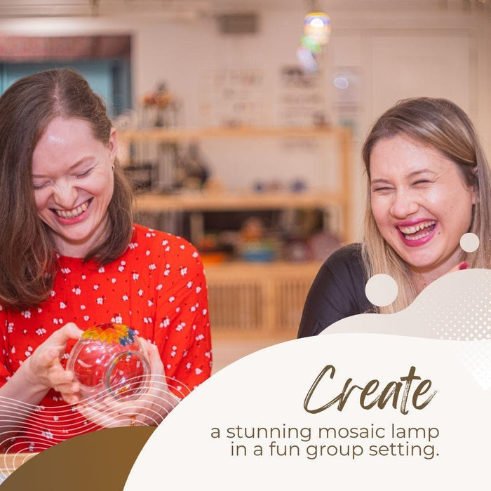 Two women laughing and holding a colorful mosaic lamp in a bright mosaic art studio
