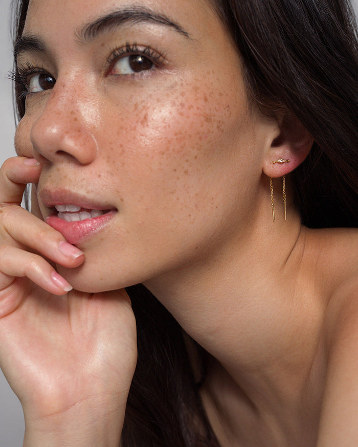A woman with long dark hair and freckled skin poses confidently with her hand near her chin, wearing the Mod + Jo Nicole Kite Threader Earring in delicate gold plating.