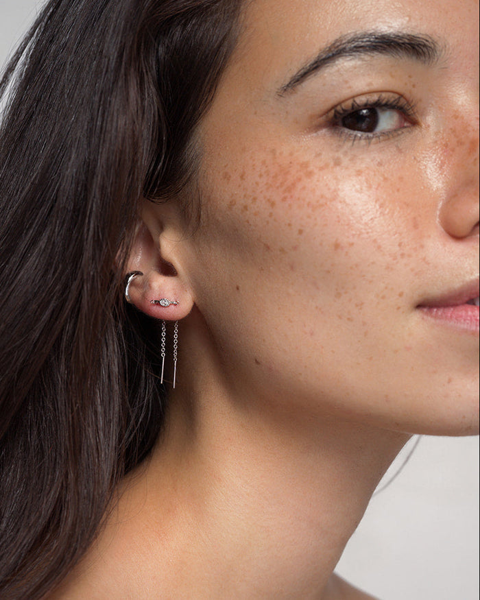 Close-up of a woman’s face, highlighting her ear adorned with three earrings: a small rhodium-plated hoop, a sparkling cubic zirconia stud, and two long Nicole Oval Threader Earrings by Mod + Jo. She has smooth, freckled skin and dark hair tucked behind her ear.