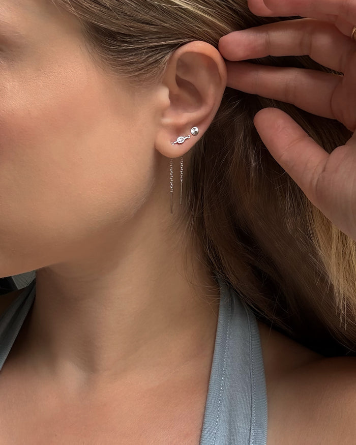 A close-up of a woman’s ear showcases three silver earrings: two cubic zirconia studs on her upper lobe and the Nicole Oval Threader Earring by Mod + Jo—a delicate, rhodium plated chain earring—on her lower lobe. She is dressed in a gray tank top and holds her hair back with one hand.