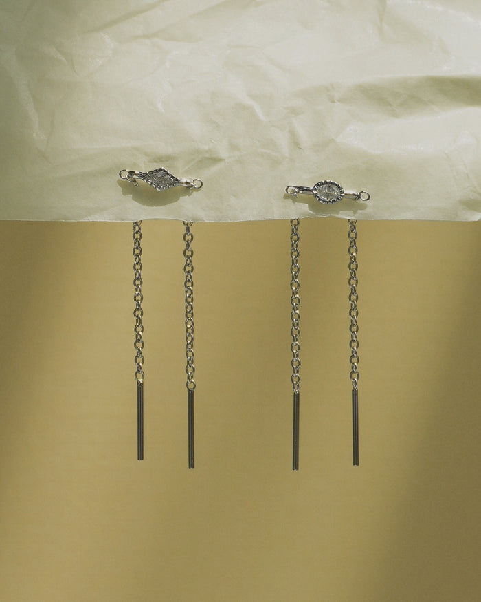 Two Nicole Kite Threader Earrings by Mod + Jo, with geometric and round tops, hang from a textured pale surface. Each earring features two delicate dangling chains ending in straight bars, set against a gold and cream background.