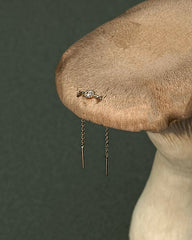 The Nicole Oval Threader Earring by Mod + Jo—a gold plated oval threader with a small round diamond and delicate dangling chains—rests on the cap of a large mushroom against a green background.